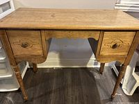 Full view of the wooden desk showing the front with two drawers and tapered legs on a dark floor.