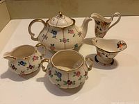 Front view of vintage cream floral ceramic teapot, two creamers with different floral patterns, and two sugar bowls with floral designs and gold trim.