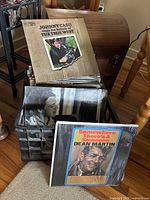 Vinyl record collection shown in metal crate with visible covers including Johnny Cash and Dean Martin albums, placed next to wooden chest and furniture.