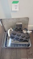 Assorted baking tools stacked on a wire cooling rack and baking trays, showing muffin pans, measuring cups, a metal box grater, sifter, and loaf pan.