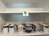 Front view of 9 assorted porcelain mugs arranged on a white shelf, showing various designs including floral, lemon motif, and colored bands with gold rims.