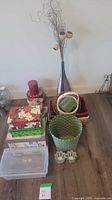 Photo showing a collection of decorative tins, woven baskets, ceramic hand figurines, a tall metal vase with twigs, and a clear plastic storage container arranged on a wooden floor.