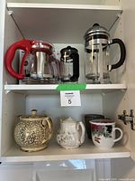 Front view of entire set in shelf with 2 shelves showing glass and ceramic cookware including teapots, French presses, and mugs