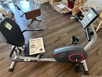 Side angle view of HealthRider H22x recumbent exercise bike showing padded seat, backrest, pedals, and part of digital console. User manual placed on the seat.
