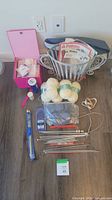 Overview of knitting supplies including metal wire basket with patterns, white yarn skeins, assorted knitting needles, and pink box with accessories.