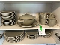 Dinnerware set stacked inside white cabinet: large plates, small plates, serving bowl with lid on large plate, and stacked cups.
