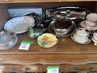 Wide view of vintage assorted dining ware and decorative items on wooden furniture including plates, cups, glass and silver plated trays and decorative figurines.