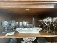 Variety of vintage glassware including etched stemmed glasses, tumblers, and a decorative porcelain platter with floral designs arranged on wooden shelf.