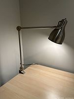Desk lamp clamped to a light wooden desk surface with articulating arm extended vertically and lampshade lit