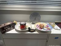 Photo of assorted decorative and collectible items on a white cabinet, including a large round metal clock, multi-colored porcelain dish with wooden mallet, carved duck figurine, glass container, and boxed kitchen gadgets.