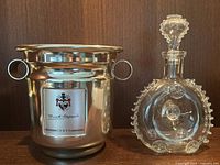 Brass champagne bucket with rings and sterling crest plate and glass Remy Martin decanter with stopper shown side by side on wooden surface against wood background.