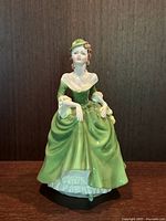 Front view of seated female figurine in green 18th century style dress, highlighting full skirt and hat.