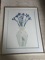 Framed print showing a vase with blue flowers and a hidden crane design in the vase, behind glass.