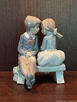 Front and side views of the LLADRO 'First Love' porcelain figurine showing seated boy and girl sharing a kiss on a bench. Glossy multicolor finish with pastel tones.