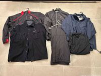 Front angled photo of all items in lot, showing jackets, windbreaker, pants, and shirt laid on floor.