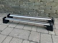 Two silver aluminum roof rack crossbars with black mounting feet for Mercedes Benz GLA 45 shown on ground against stone wall.