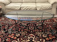 Front view of oval glass coffee table with metal curved legs and copper-tone painted accents, placed on a patterned rug in front of beige sofa.