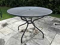 Top view of round metal mesh patio table showing umbrella hole at center, black curved legs, outdoors on stone patio.