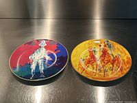 Two LeRoy Neiman limited edition painted Royal Doulton collector plates displayed side by side on reflective surface, one with bright colorful Pierrot image and the other with yellow background horse racing scene.