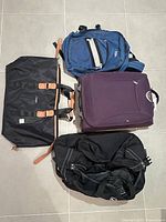 Top-down view of four travel bags on floor: vintage blue MEC backpack, purple Jaguar carry-on suitcase, black Bugatti duffle, black Puma duffle.