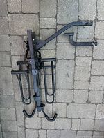 Complete black metal trailer hitch bike rack with angled arm and two U-shaped wheel hooks laid flat on stone pavement.