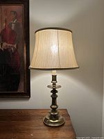 Table lamp lit on wooden surface beside framed painting, showing full lamp with shade and turned brass base.