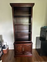Wooden two-piece bookshelf with three shelves and a cabinet with brass handles, shown standing against a wall on hardwood floor.