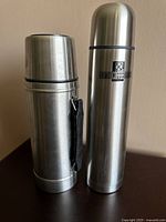 Two stainless steel thermos flasks standing side by side, showing their exterior, lids and attached handle on shorter flask.