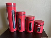 Four red glass and plastic canisters arranged in descending height order, showing vertical glass windows and screw-on lids.