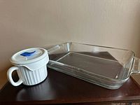 Clear rectangular glass baking dish with integrated glass handles, placed on a surface beside a white ceramic mug with a lid.