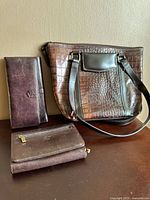 Photo of brown crocodile-textured leather handbag with shoulder strap and two brown leather wallets on a beige surface.