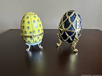 Two decorative eggs standing upright: one dark blue with gold design and legs, one yellow with blue flowers and gold trim.