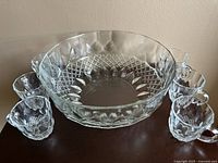 Wide shot of the clear punch bowl surrounded by six matching cups, showing the diamond cut pattern and handles on bowl and cups.