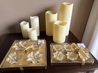 Photo of boxed votive wax candle sets with gold floral motifs and five battery operated candles of various heights on table.
