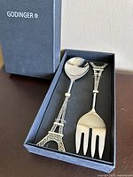 Set of two serving utensils in a black box with Godinger branding. Spoon and fork with detailed Eiffel Tower shaped handles made of metal with nickel finish.