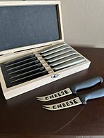 Wooden box containing six steak knives with stainless steel handles and blades, displayed with two cheese knives with black plastic handles and 'CHEESE' cut-out blades.