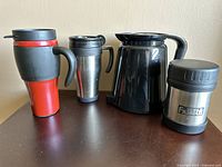 Front view of four drinkware items: a red and black travel mug, a stainless steel travel mug, a black Keurig-style coffee carafe, and a small stainless steel thermos.