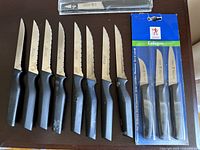 View of eight J.A. Henckels serrated steak knives with black ergonomic handles laid out side by side, showing the blades and minor wear on handles