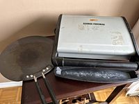George Foreman electric grill closed position, showing wear and residue marks on silver lid and black base, with tava pan handle visible.