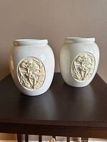 Two off-white ceramic vases with crackled glaze and oval bird floral relief, shown side by side on a wooden surface.