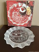 Photo of Mikasa 9 inch engraved glass holiday plate with box in background, showing scalloped frosted edges and holiday scene engraving.