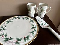 Photo showing two large dinner plates, two mugs, and one cake server with holly and ribbon pattern on white bone china with gold edges.