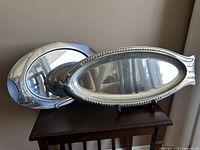 Photo showing two oval silver stainless steel serving platters stacked on a stand on a wooden table near a window.