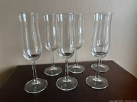 Six clear glass champagne flutes arranged on a dark wood surface, front view showing tall slender glasses.