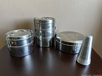 All four items displayed on a flat surface including two tiffin carriers, one ventilation container with perforated lid, and one small cone-shaped item