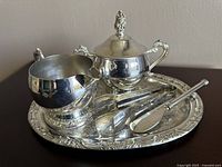 Photo showing silver-plated creamer, lidded sugar bowl, serving tongs, and butter knife arranged on the engraved platter.