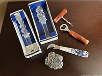 All items in the wine tools set displayed on a table, with two crystal bottle stoppers in boxes, corkscrew and bottle opener with orange marble handles, and a porcelain-handled foil cutter.