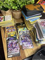 Multiple stacks of unsleeved and sleeved Pokémon cards on wooden surface, with plant pots in background