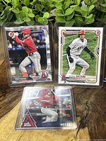 Three Shohei Ohtani baseball cards in protective sleeves on a wooden surface with green plants in the background.