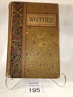 Front cover of the 1892 hardcover book 'The Poetical Works of John Greenleaf Whittier' showing elaborate gold and black floral design with red details.